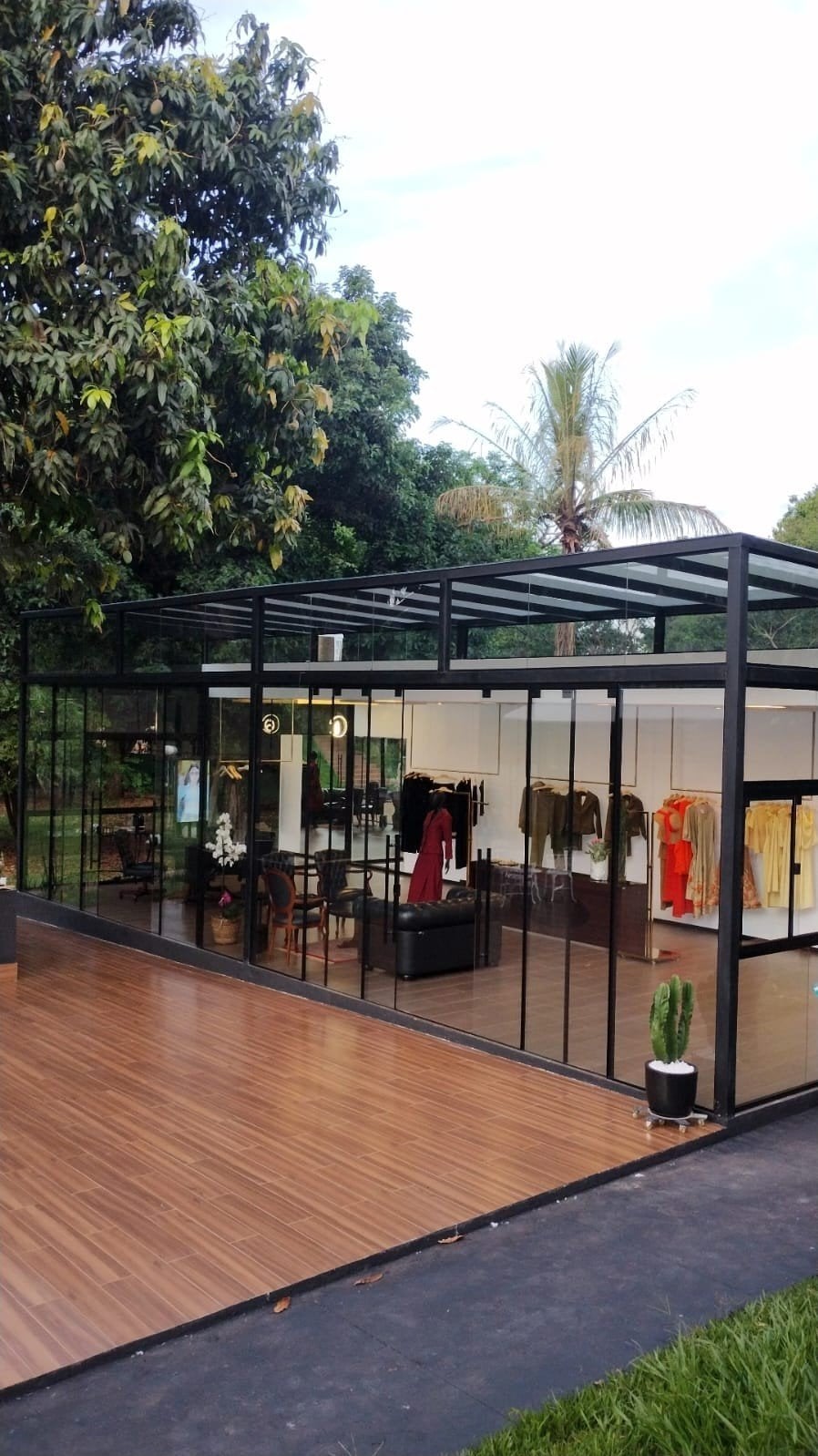 Fachada de vidro do Louback Atelier cercada de verde, com araras de roupas no interior